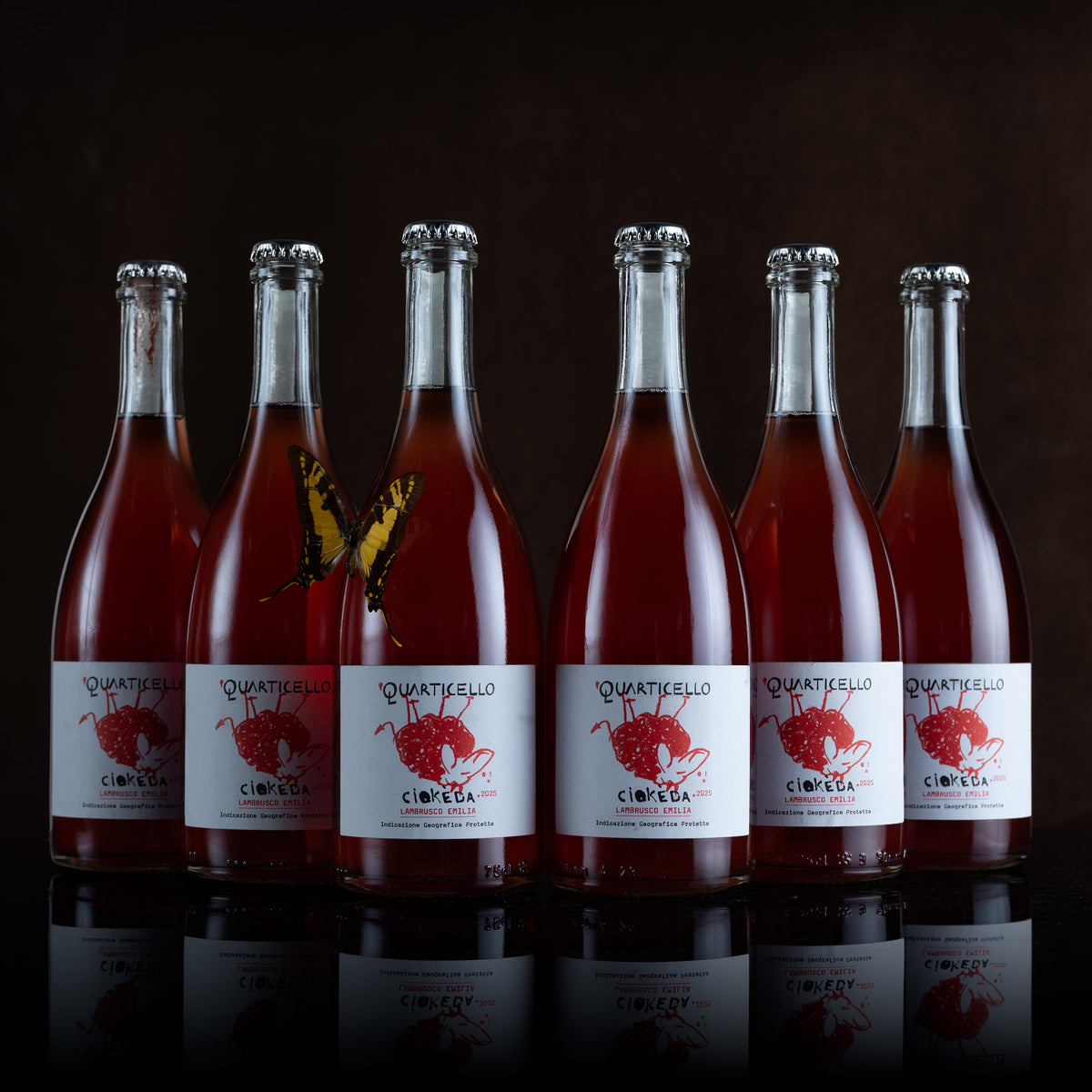 Six glass bottles of Quartesello Cimonca liqueur with deep red liquid, lined up on a reflective dark surface against a black background. A yellow-and-black butterfly is perched on the second bottle.