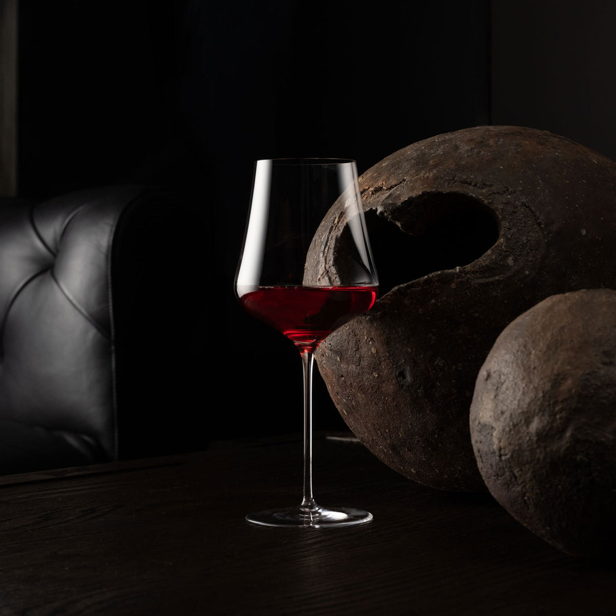 A clear stemmed wine glass with a flared bowl, partially filled with red wine, standing on a dark wooden table beside two large textured brown stone spheres and a black leather chair in a dim interior setting.