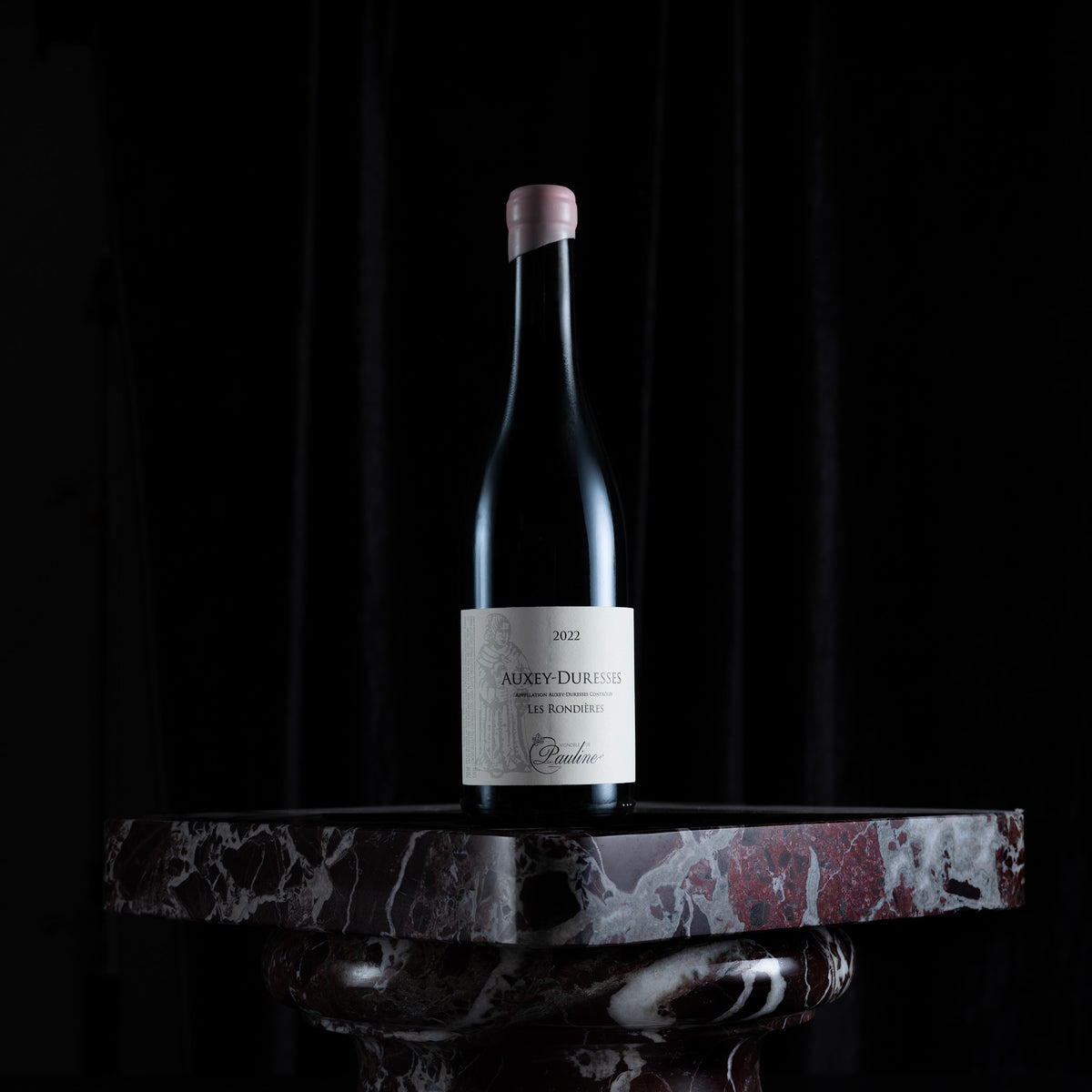 A single dark wine bottle with a pale pink capsule and a white label reading Audrey Duchamp, displayed on a polished red-and-white marble pedestal against a black background.