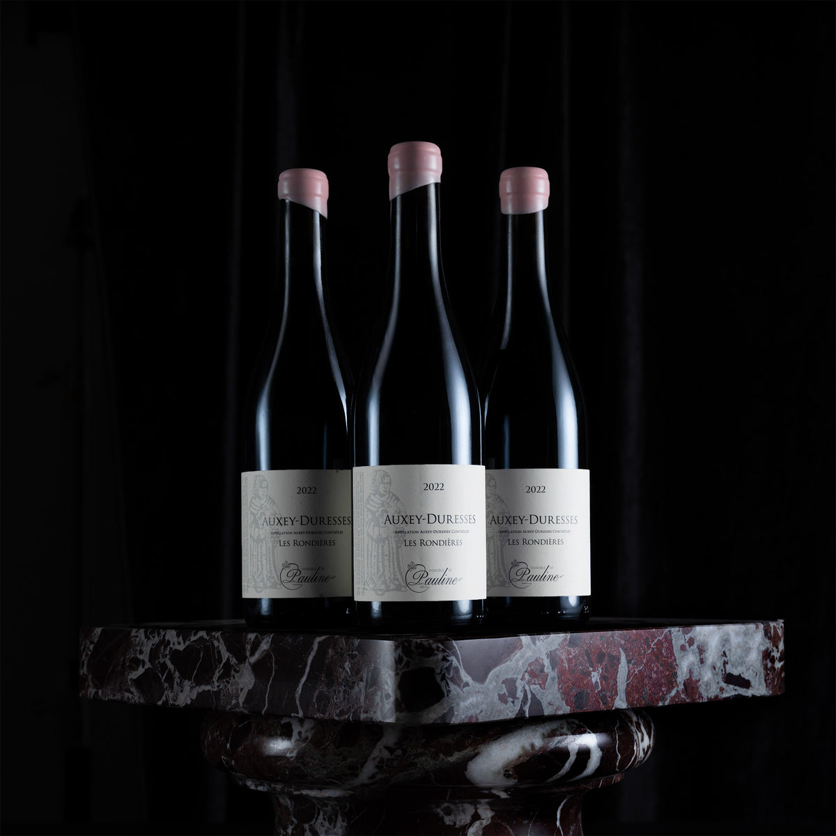 Three dark glass wine bottles with pale pink capsules and white labels reading Auxey-Durier, displayed on a polished red-and-white marble pedestal against a black background.