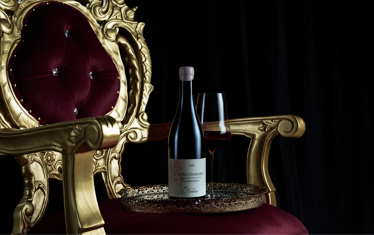 A bottle of red wine and a filled wine glass sit on a round tray placed on the arm of an ornate gold chair with deep burgundy velvet upholstery, set against a black background.