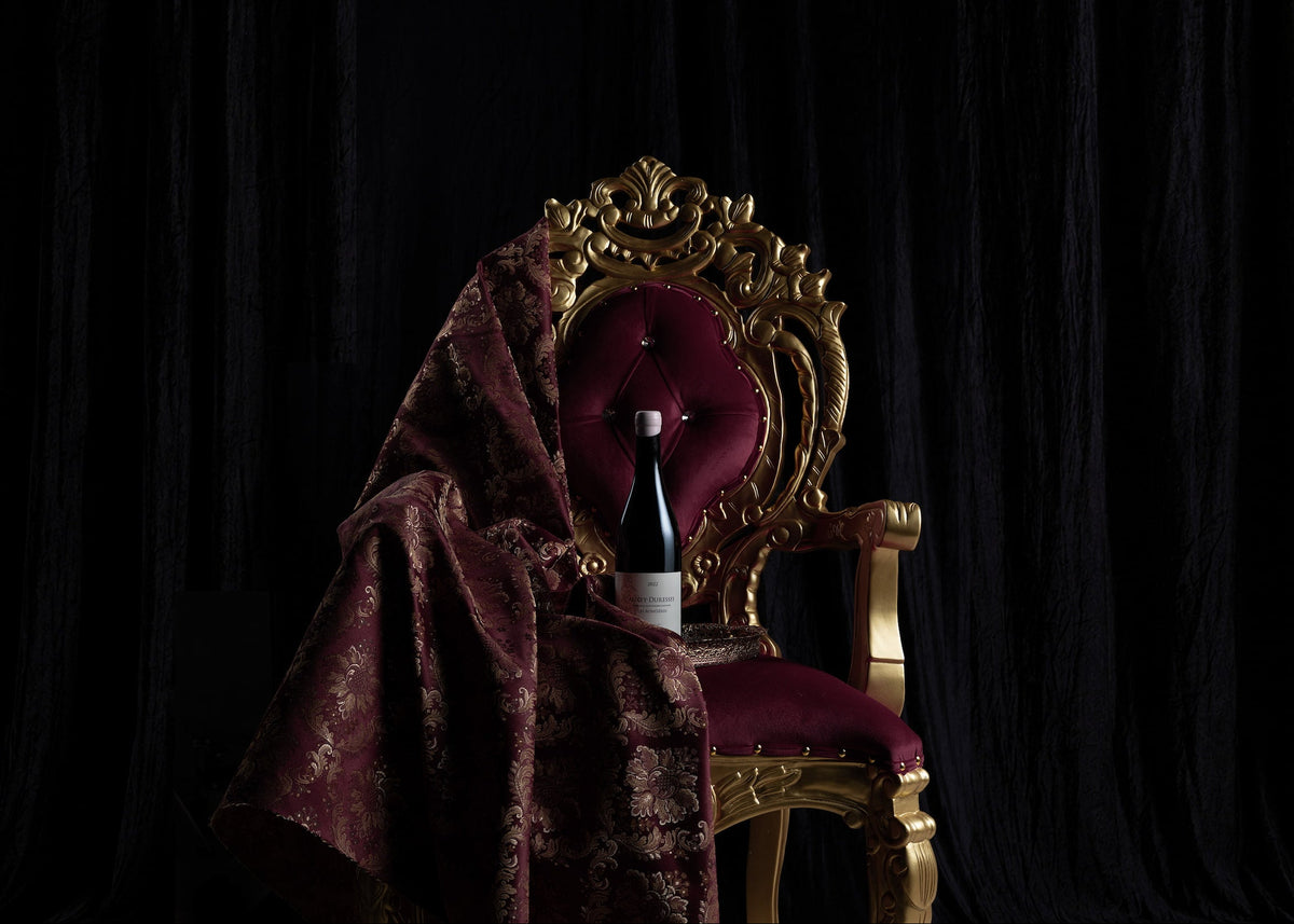An ornate gold throne chair with deep burgundy velvet upholstery, draped with a matching burgundy damask fabric, with a dark wine bottle resting on the seat, set against a black curtain backdrop.