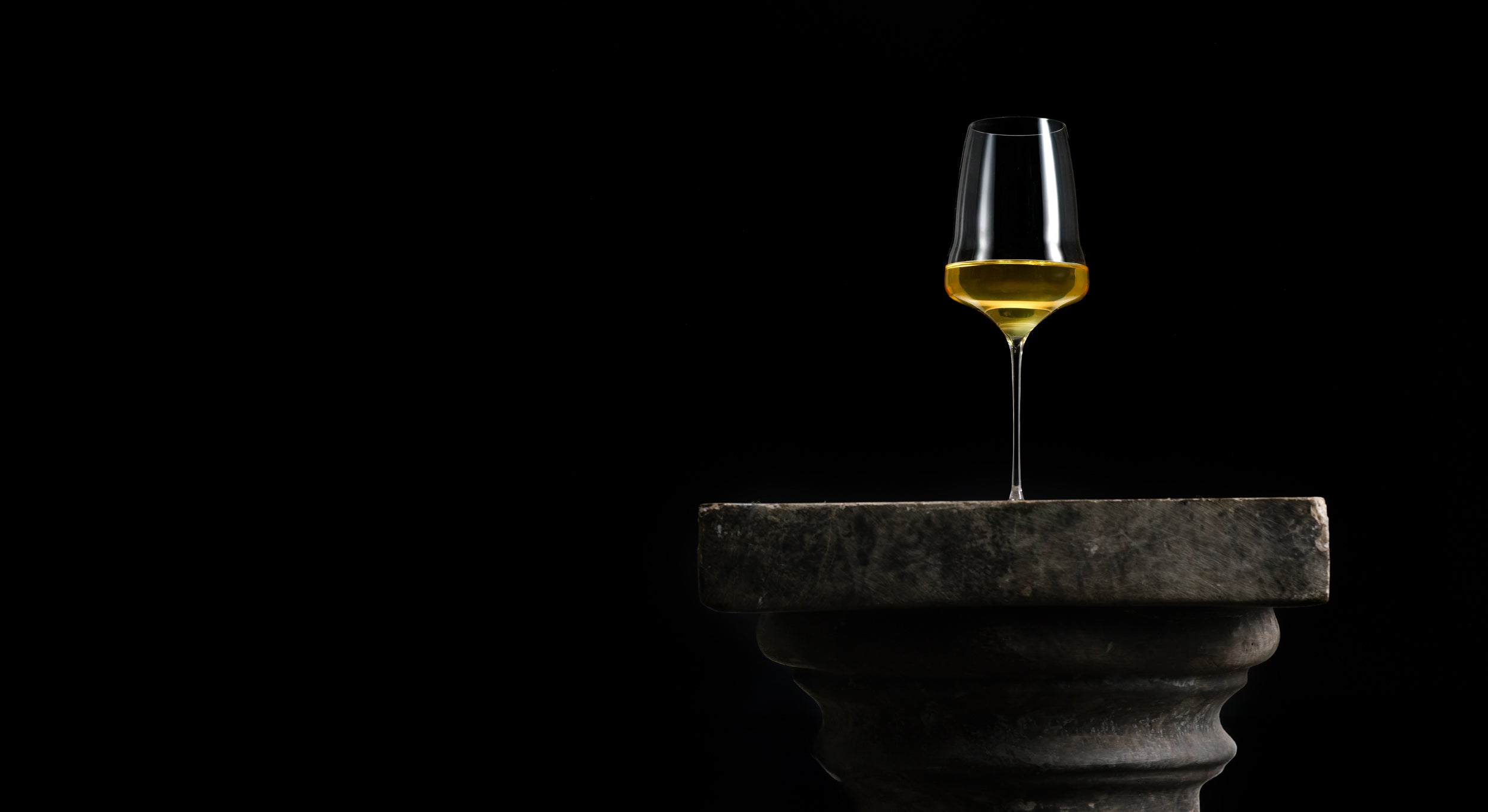 A clear wine glass containing a golden yellow liquid, balanced on the edge of a dark stone pedestal against a solid black background.