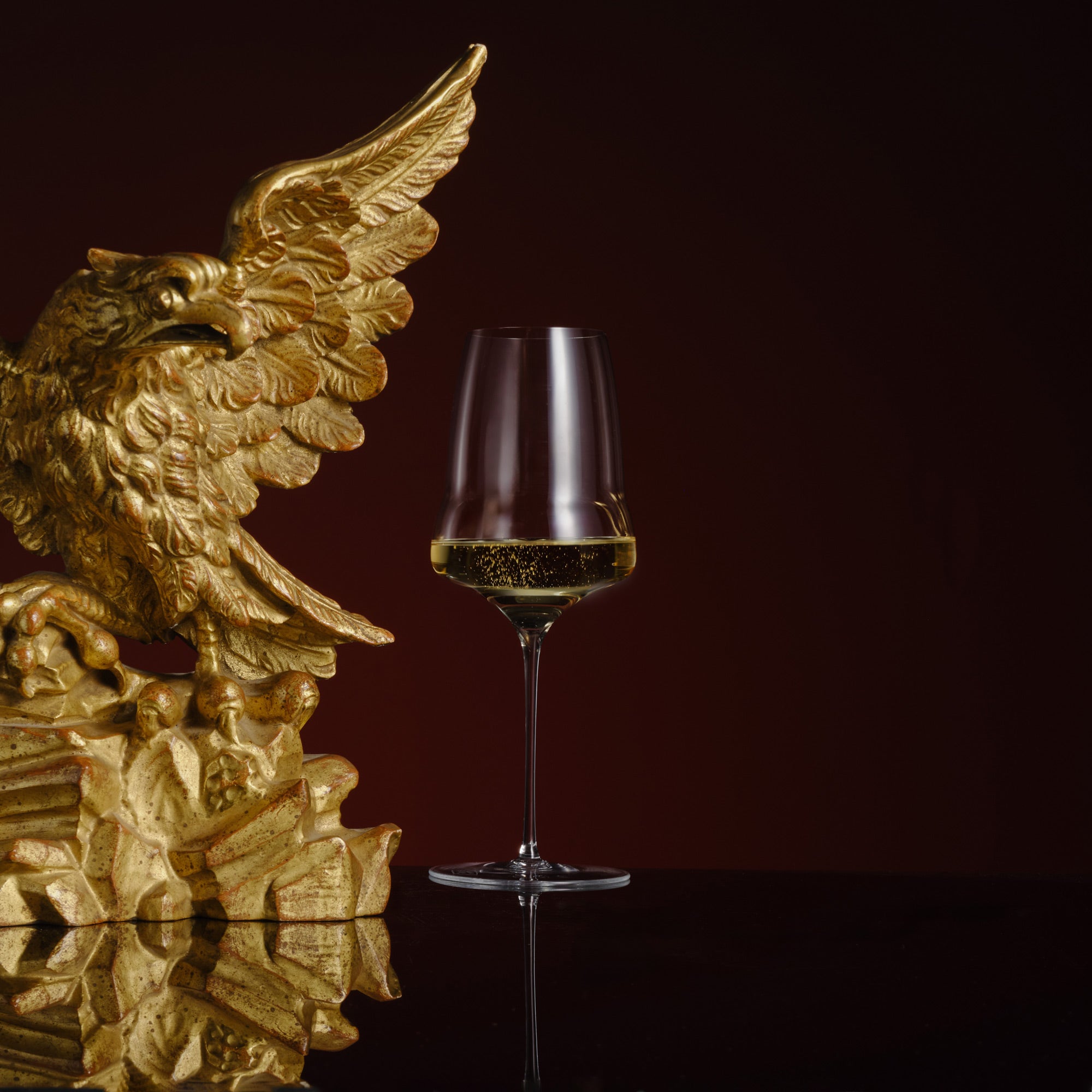 Gold eagle head sculpture next to a glass of white wine on a dark background