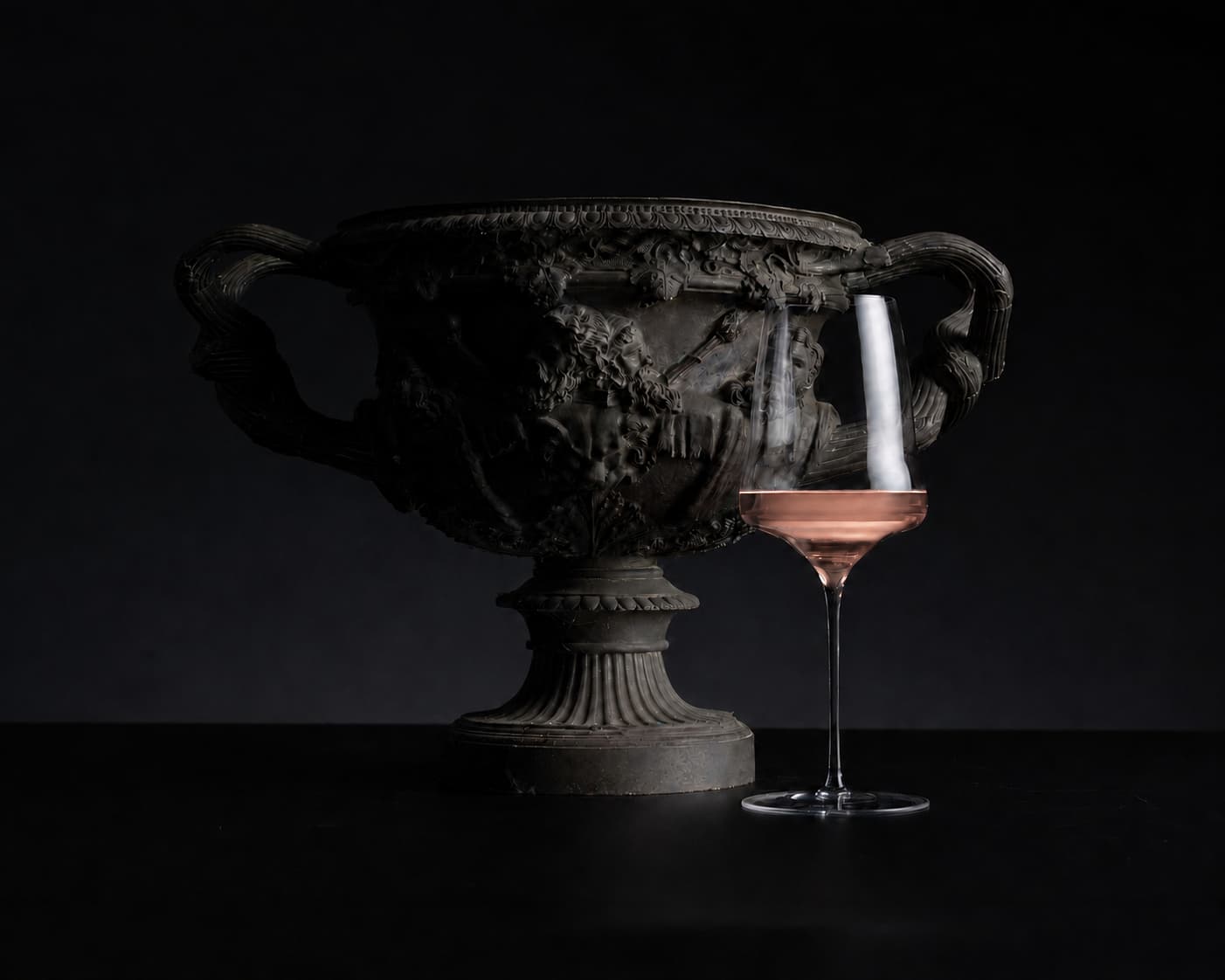 An ornate dark bronze classical urn with sculpted details and two handles, displayed next to a coupe glass containing a pinkish liquid, set against a solid black background.