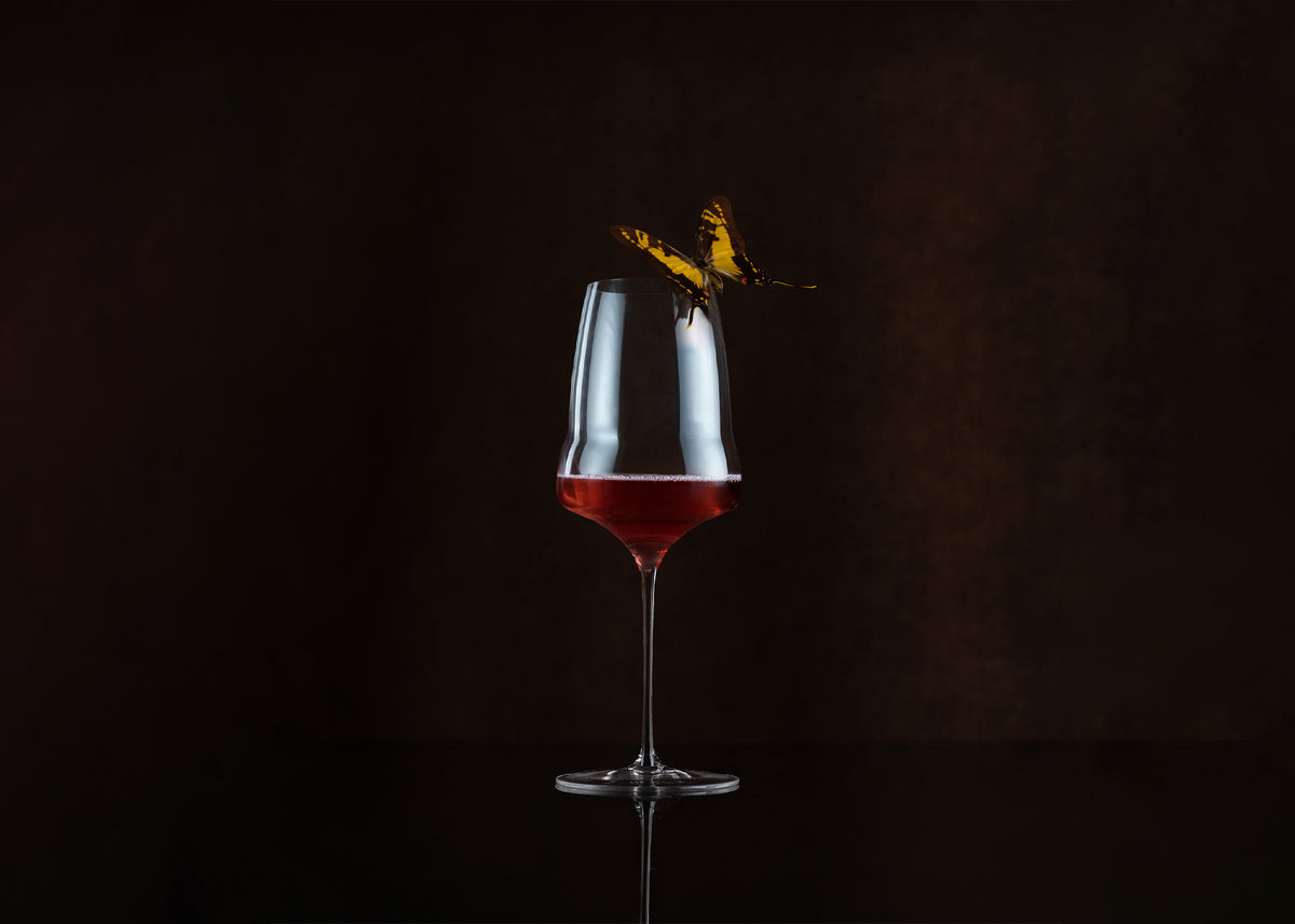 A clear stemmed wine glass with a wide bowl, partially filled with red wine, set against a dark brown background. A small yellow-and-black butterfly is perched on the rim of the glass.