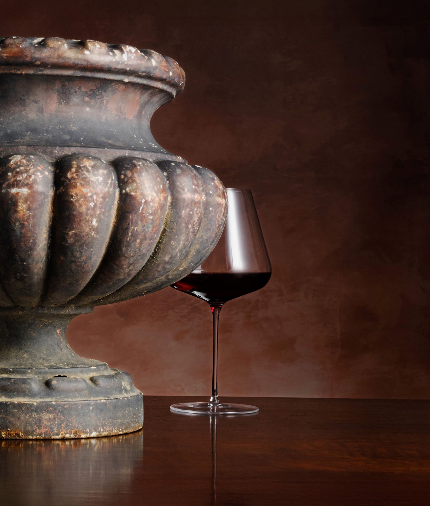 Wine glass with red wine next to an ornate dark stone urn on a wooden surface.