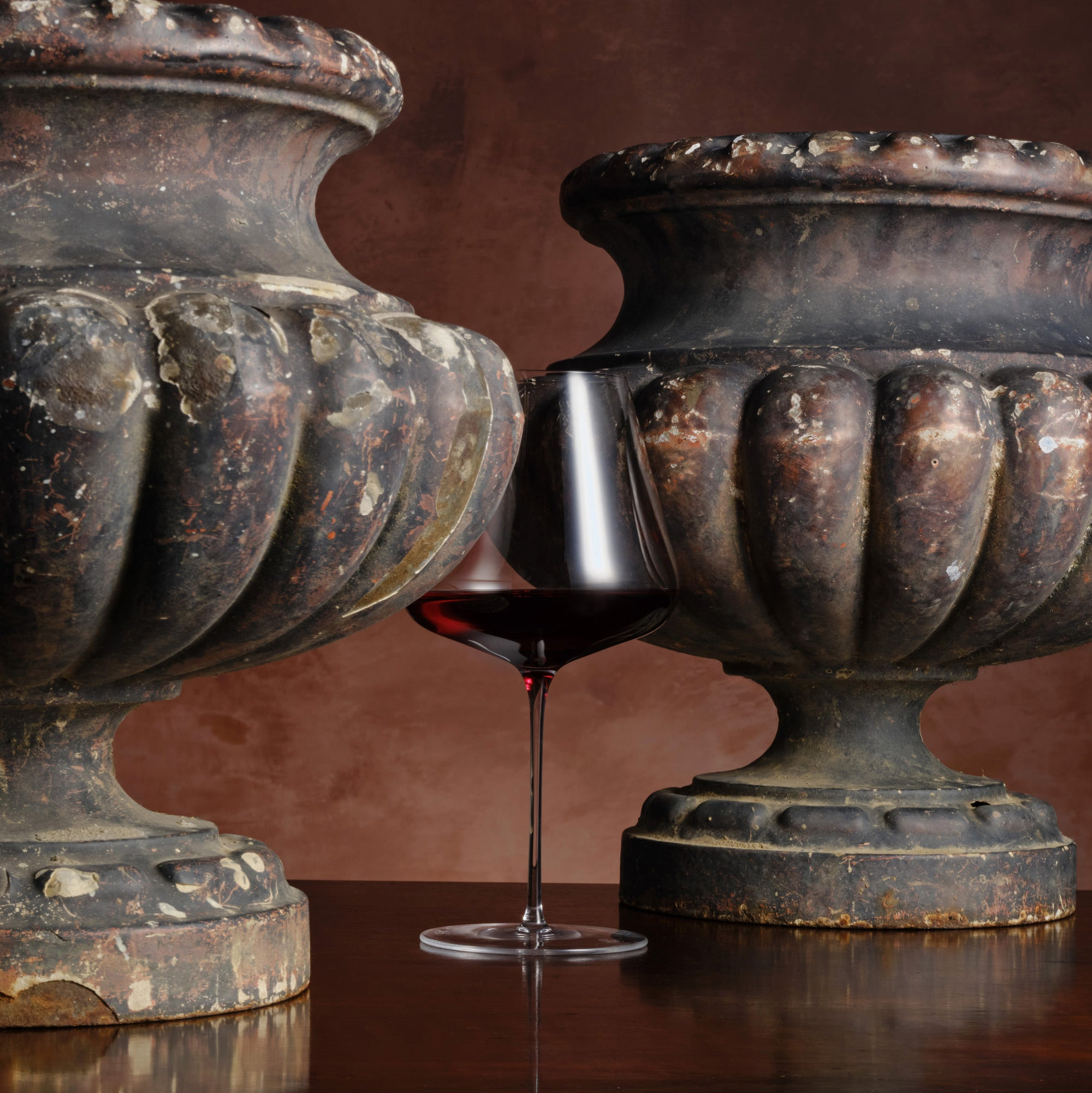 Wine glass with red wine between two rustic stone urns on a reflective surface.