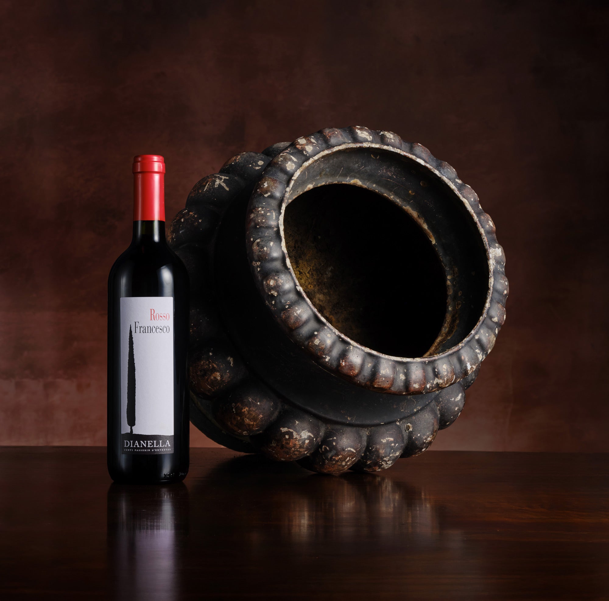 Wine bottle with red cap and label next to an ornate dark vase on a reflective surface.