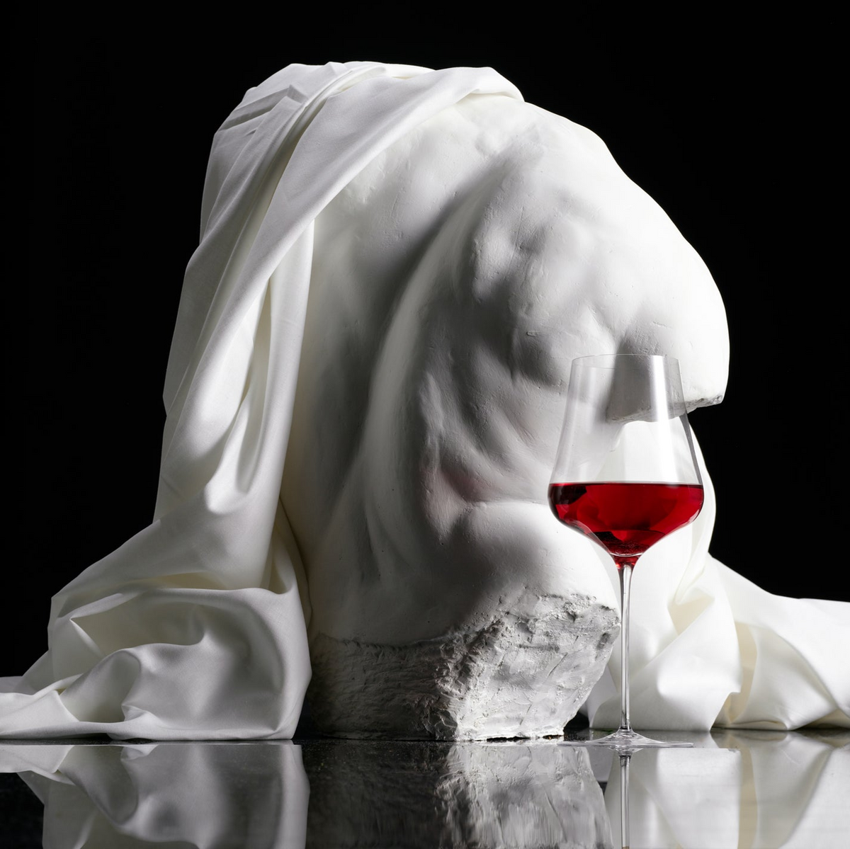 Marble bust draped with a white cloth next to a glass of red wine on a reflective surface.