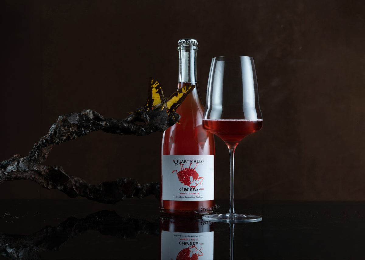 A bottle of red wine with a butterfly on the label stands beside a filled wine glass on a dark reflective surface against a dark brown background.
