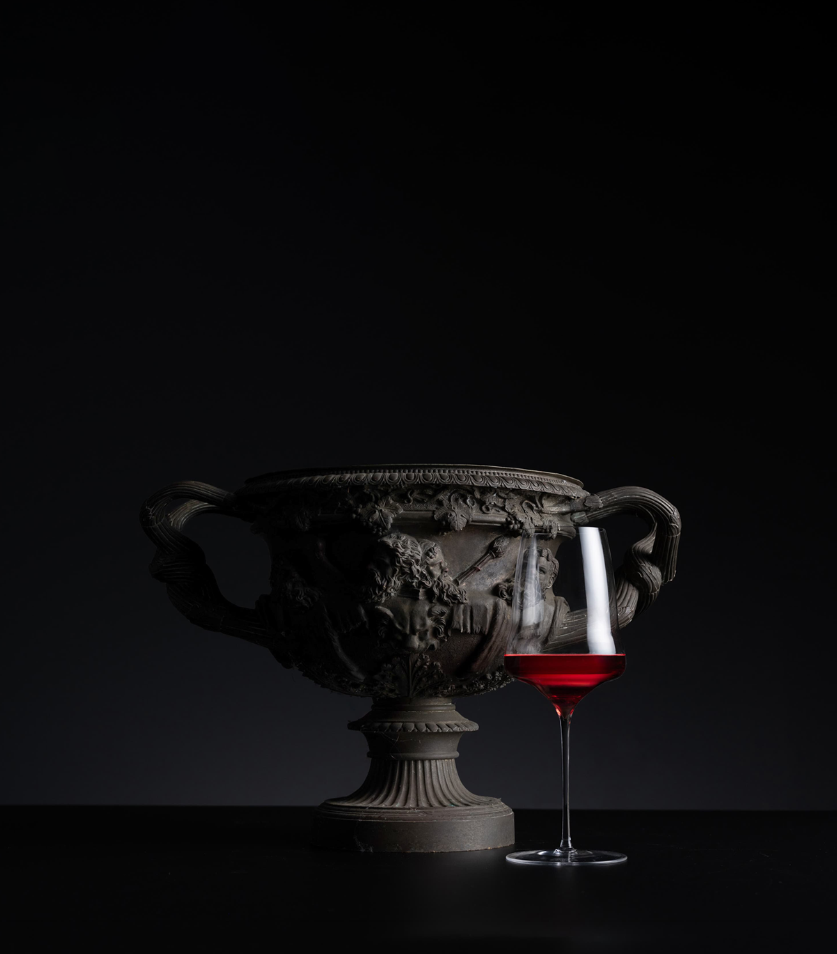 An ornate dark gray carved stone or metal urn with two handles sits on a black surface against a black background, next to a clear wine glass filled with red liquid.