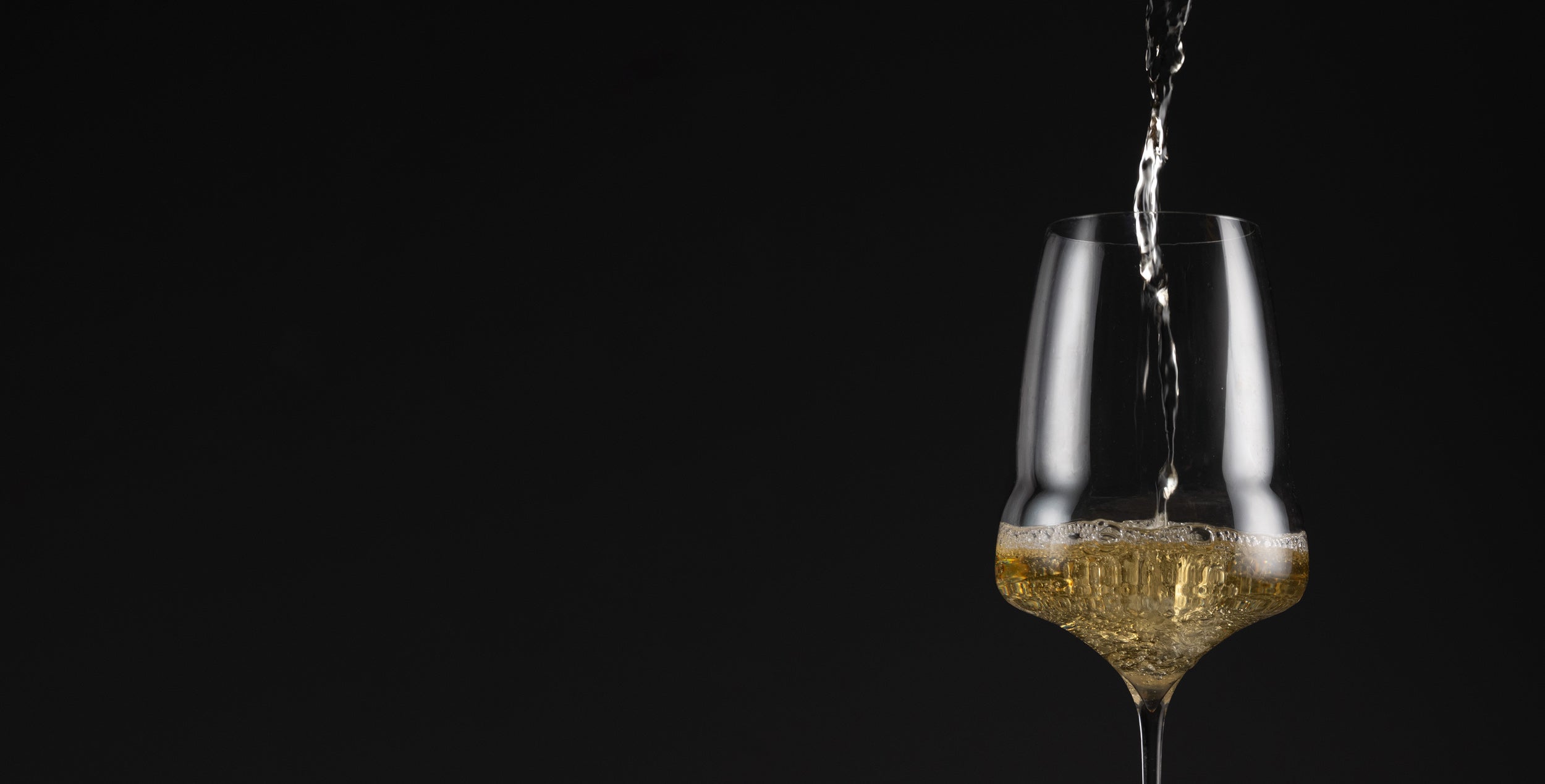 A clear wine glass on a black background with a stream of golden liquid being poured into it, creating visible bubbles.